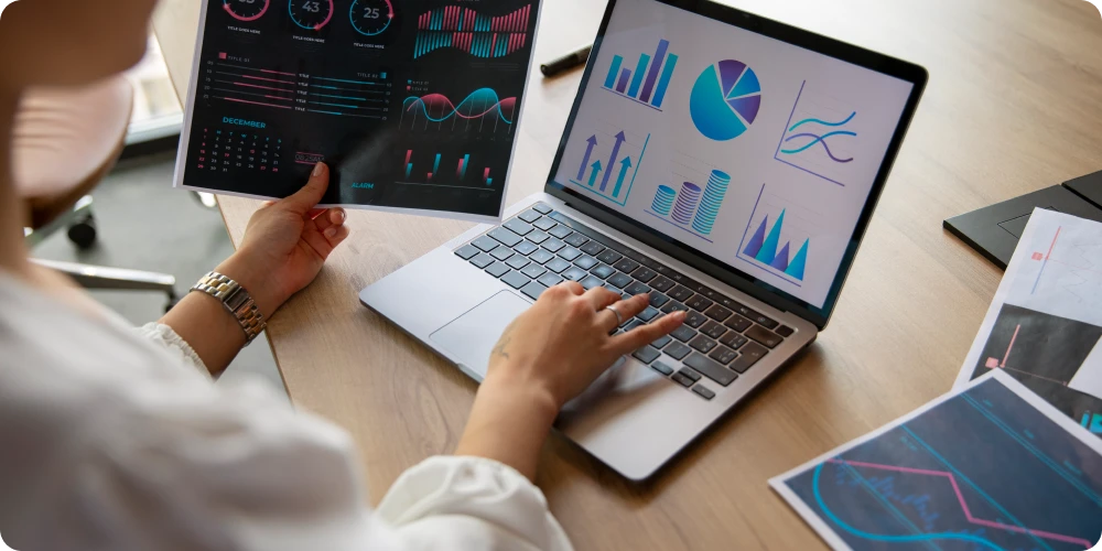 Person analyzing data on a laptop with charts and graphs, holding a printed sheet showing data visualizations on a desk.